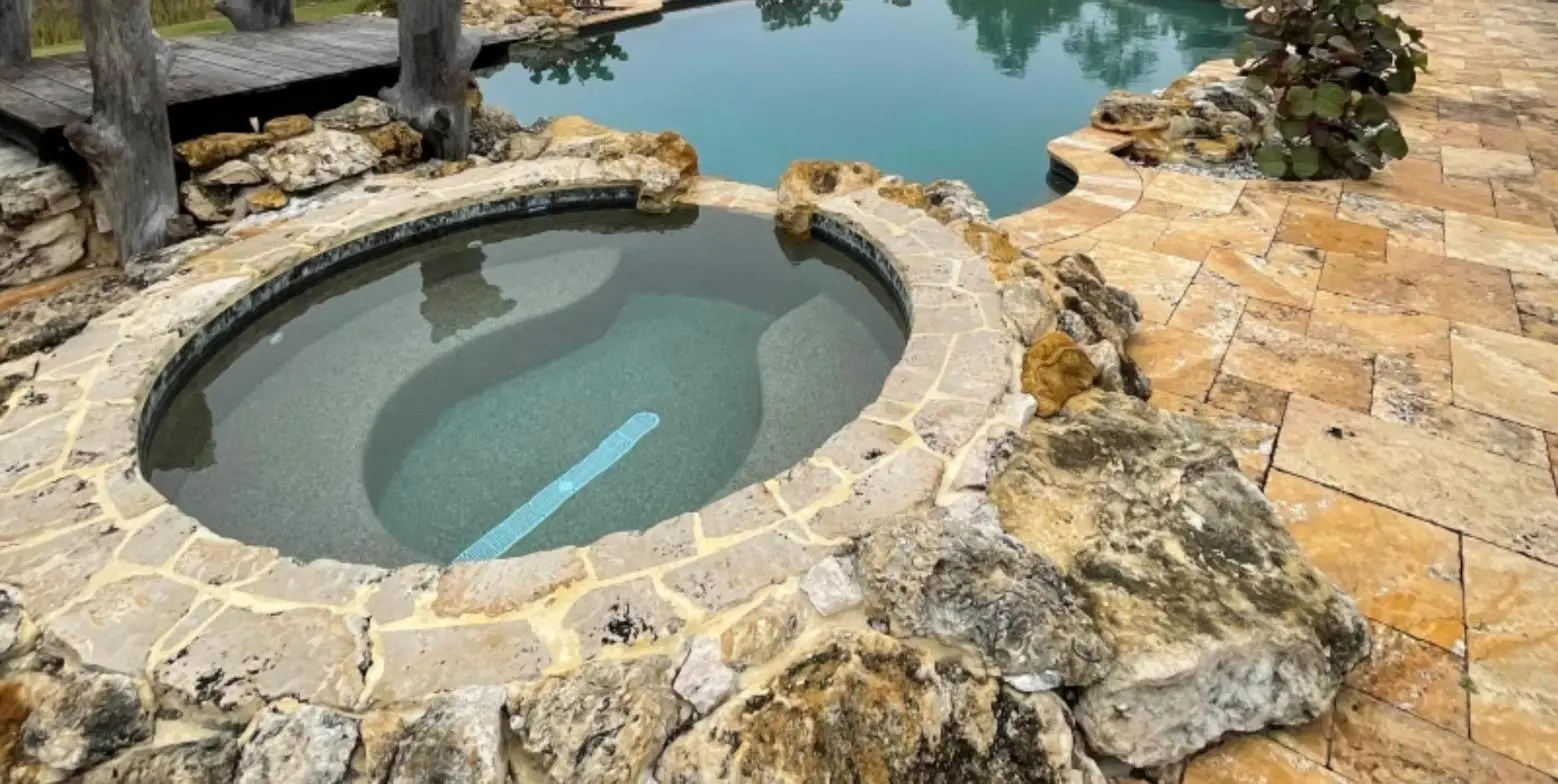 Round spa with a natural stone edge beside a freeform pool and travertine pavers